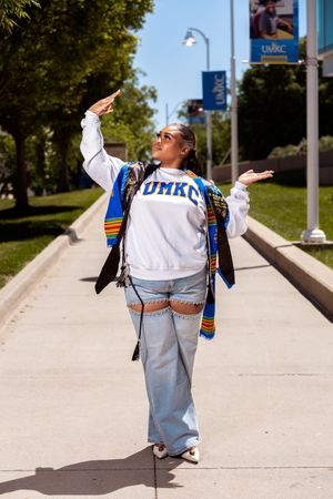 A person in a white Drake University sweatshirt and jeans strikes playful poses on a campus sidewalk on a sunny day.