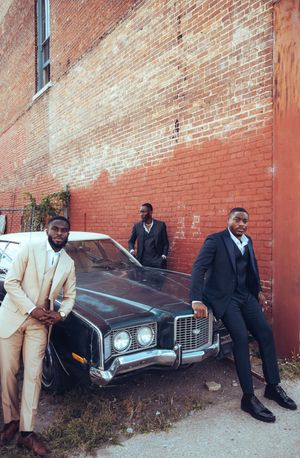 Group of people in formal attire sitting and standing against red brick wall next to vintage car.