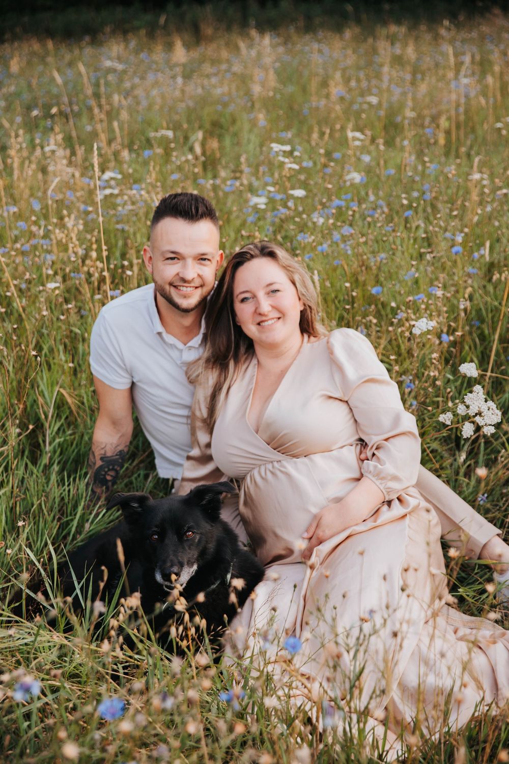 A couple sits in a wildflower meadow with their black dog on a sunny afternoon.