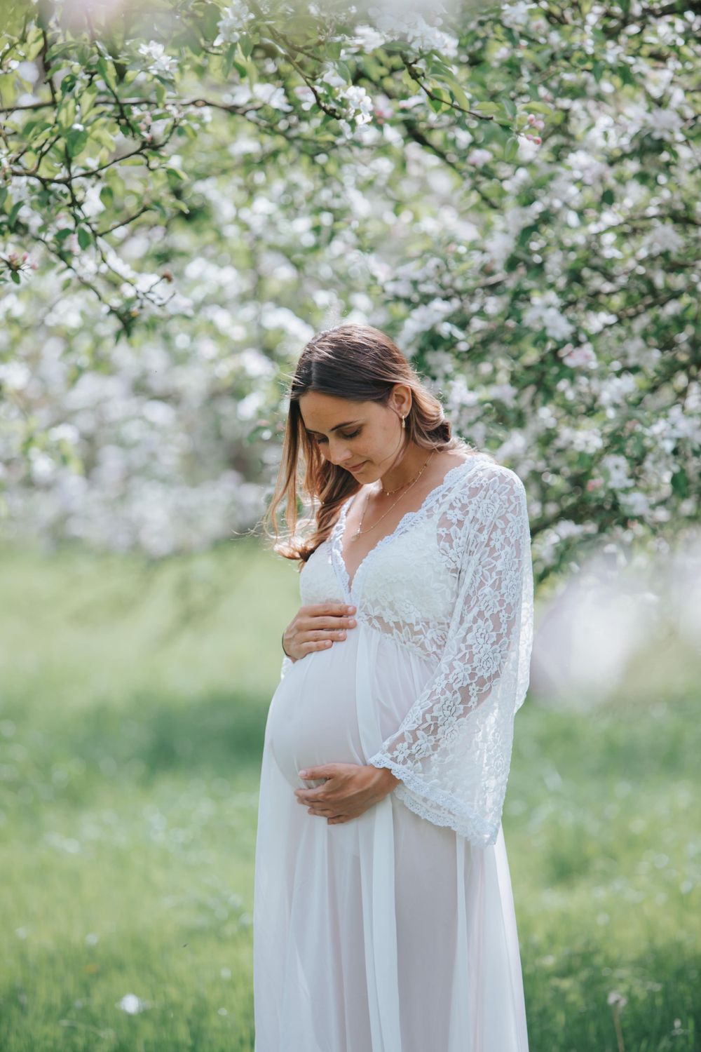 A maternity photo of an expectant mother in a white lace dress among blooming trees.