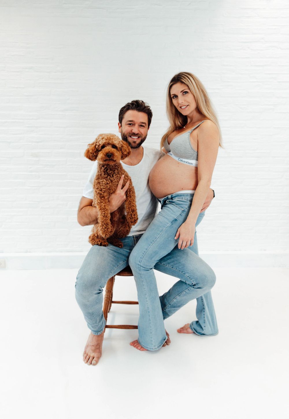A couple sits on a wooden stool with their brown goldendoodle dog in a bright white studio setting.