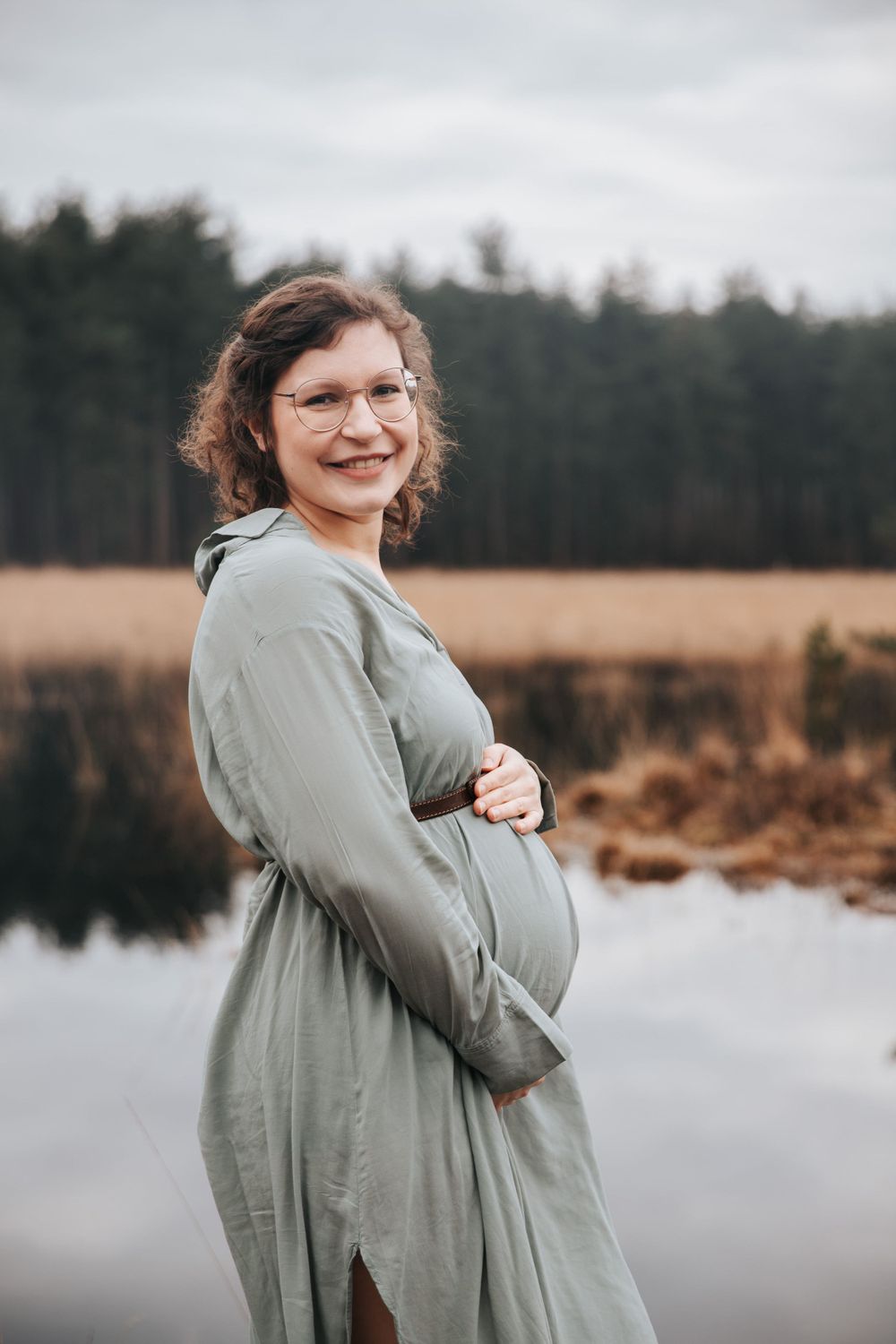 Maternity photo in sage green dress against moody lake backdrop.