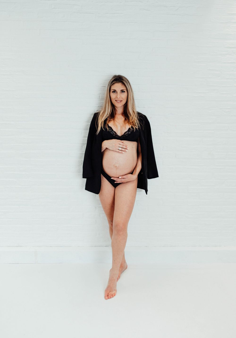 Artistic maternity photo of an expectant mother wearing a black cardigan against a white wall in minimal style.