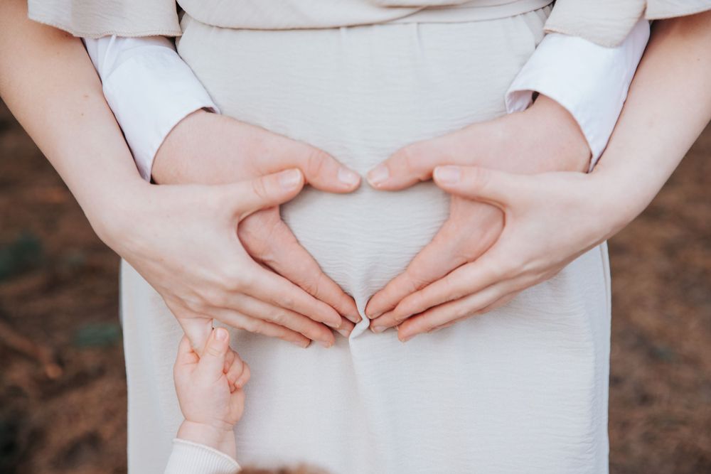 Hands forming heart shape over pregnant belly in white dress.