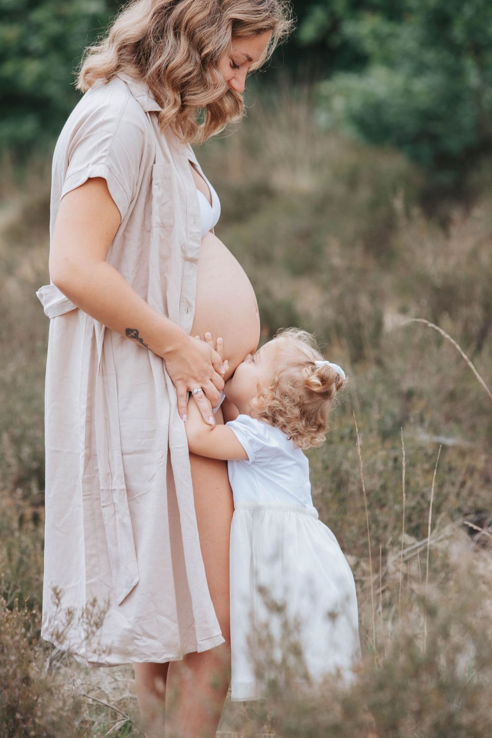 Sweet maternity moment captured outdoors in a field with a light beige dress and soft natural lighting.