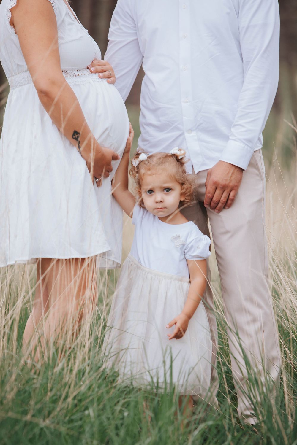 A family wearing white stands together in tall grass, with a toddler in a tulle dress between the parents.