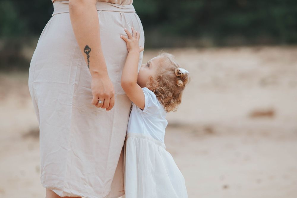 Parent and child wearing light colored dresses embrace in tender moment on sandy beach at sunset.