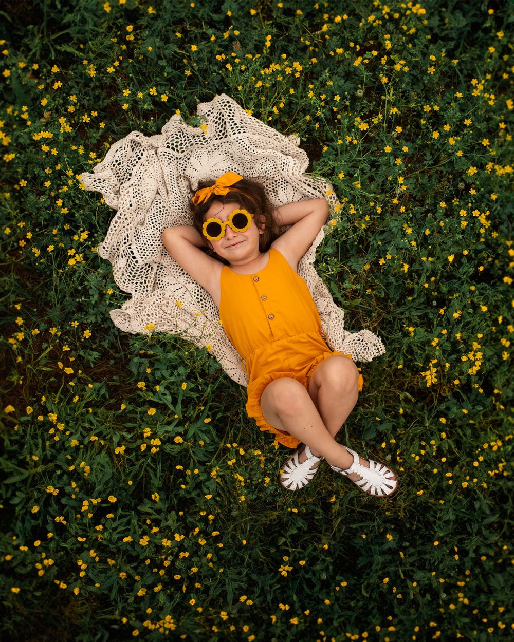 A little girl wearing an orange dress lies on grass surrounded by small yellow wildflowers on a sunny day.