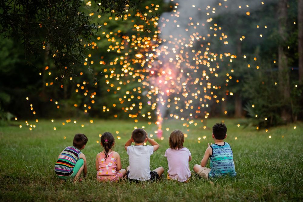 A group of kids sits on a grassy hill watching fireworks light up the evening sky with golden sparkles in the background.