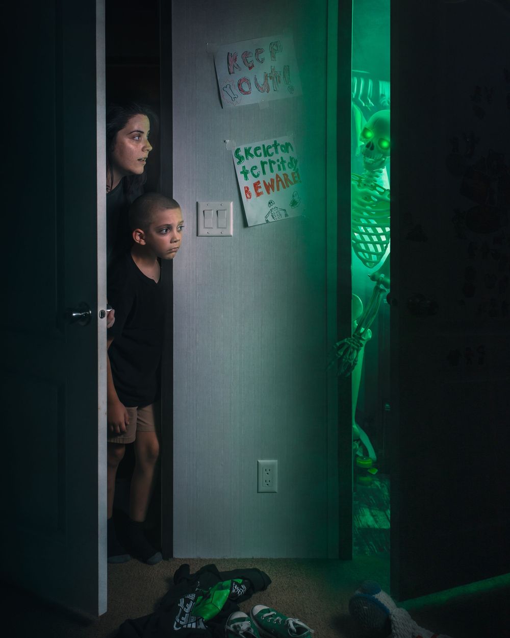 Two figures peer around a doorway into a dark room illuminated by eerie green light creating a mysterious atmosphere.