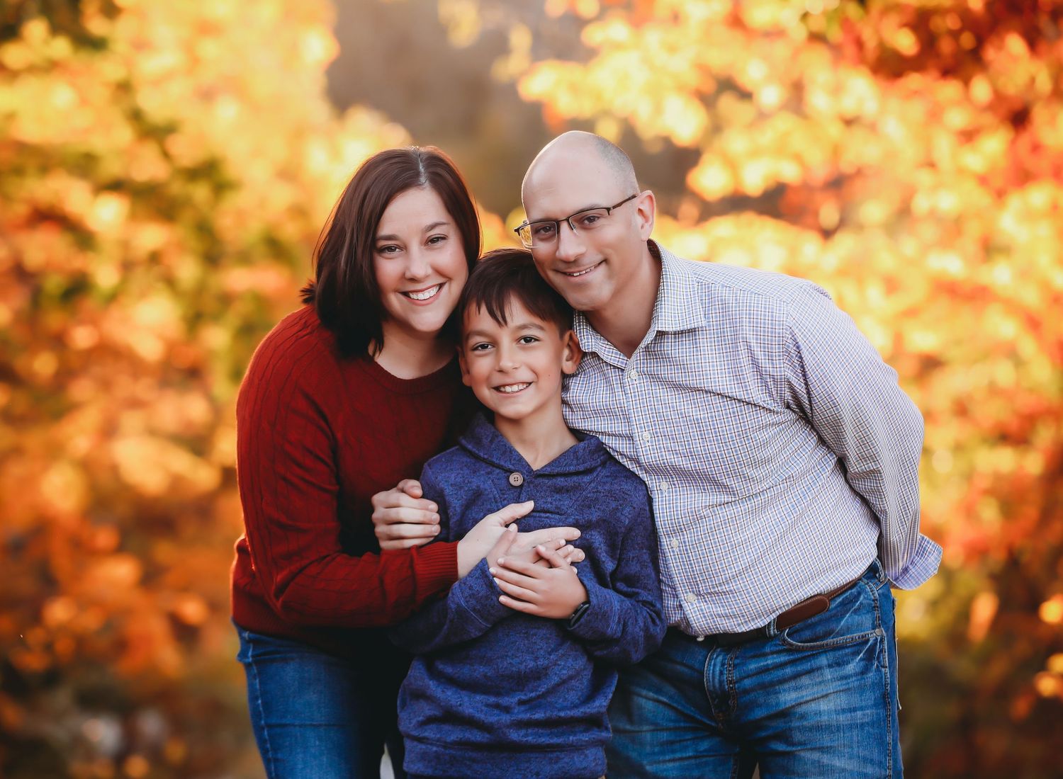A family of three poses together outdoors against vibrant orange autumn foliage in the background.