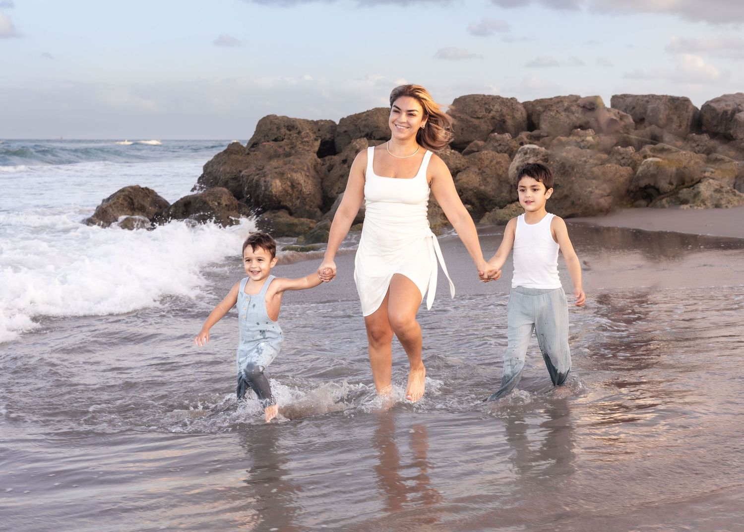 A mom runs with her sons in the ocean waves along a rocky beach shore at sunset.
