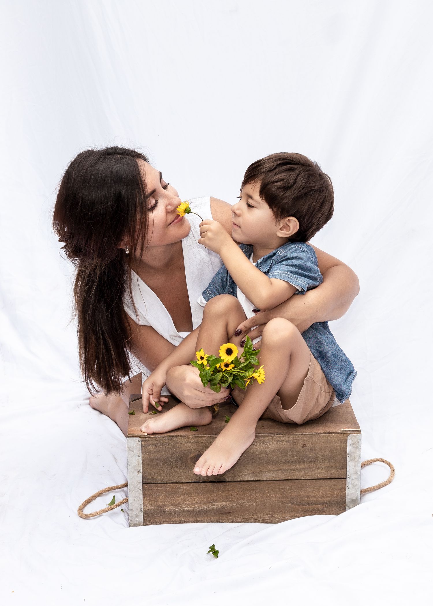 A parent and child share playful moments on a wooden box during a white-sheet mini photo session.