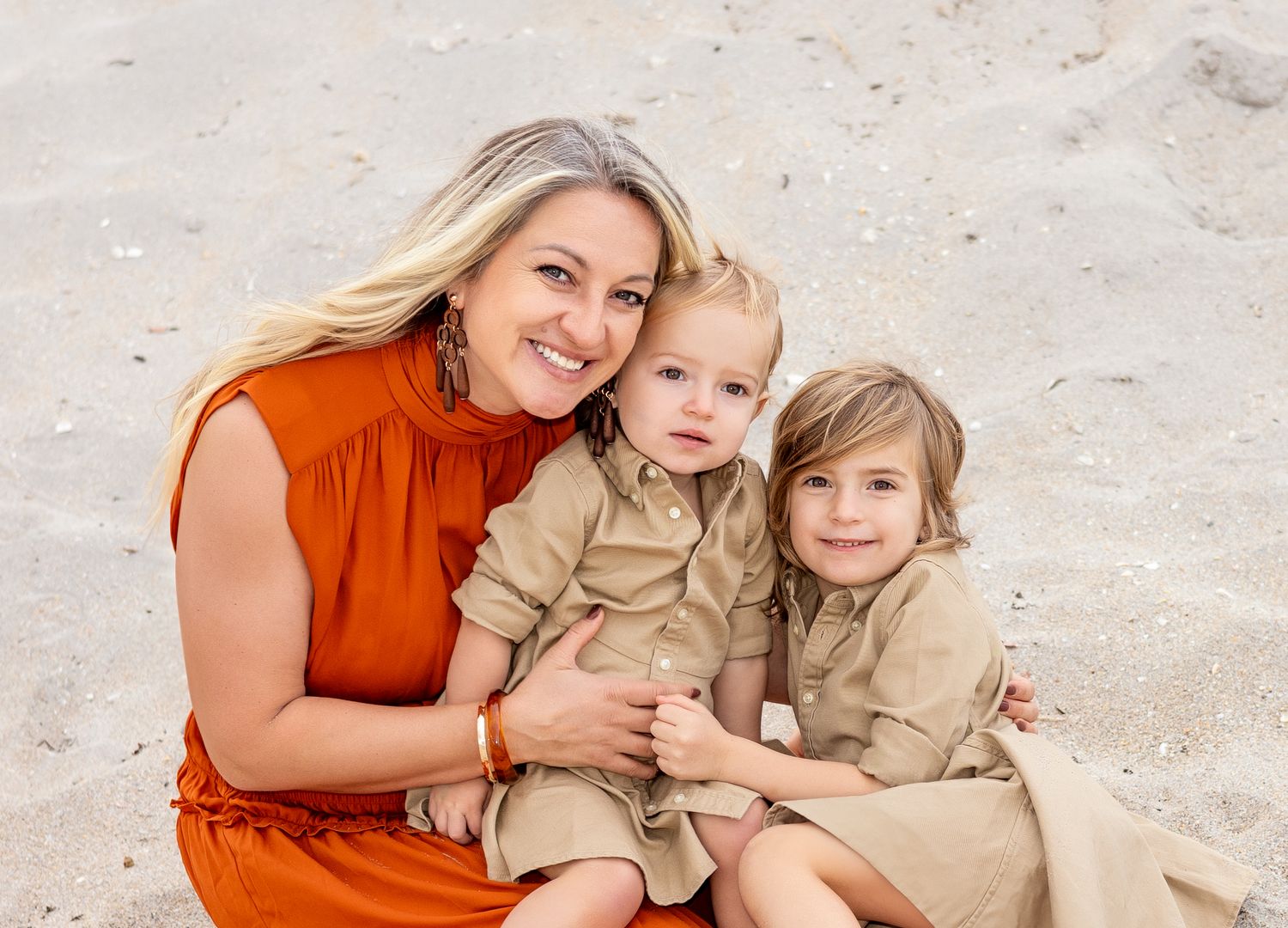 A mother in a rust-colored dress embraces her children on a sandy beach during a heartfelt photo session.