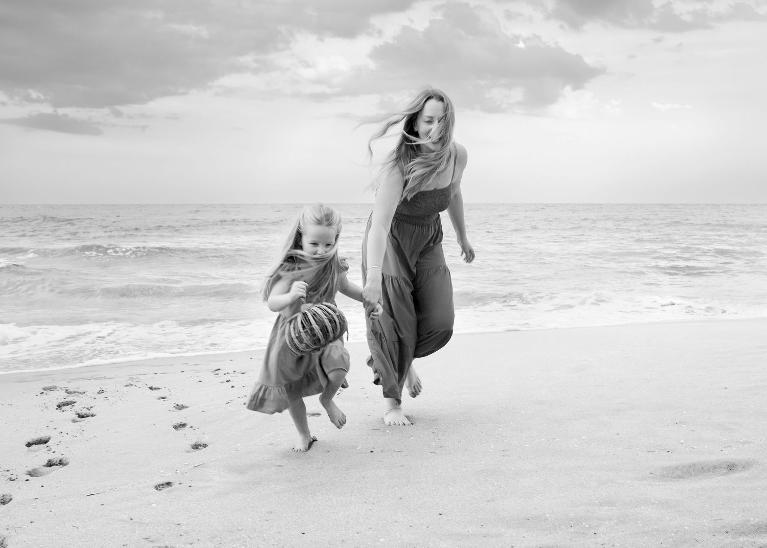 A black and white photo shows mom and daughter joyfully running along a beach with windswept hair and long dresses. Bohemian style dresses.