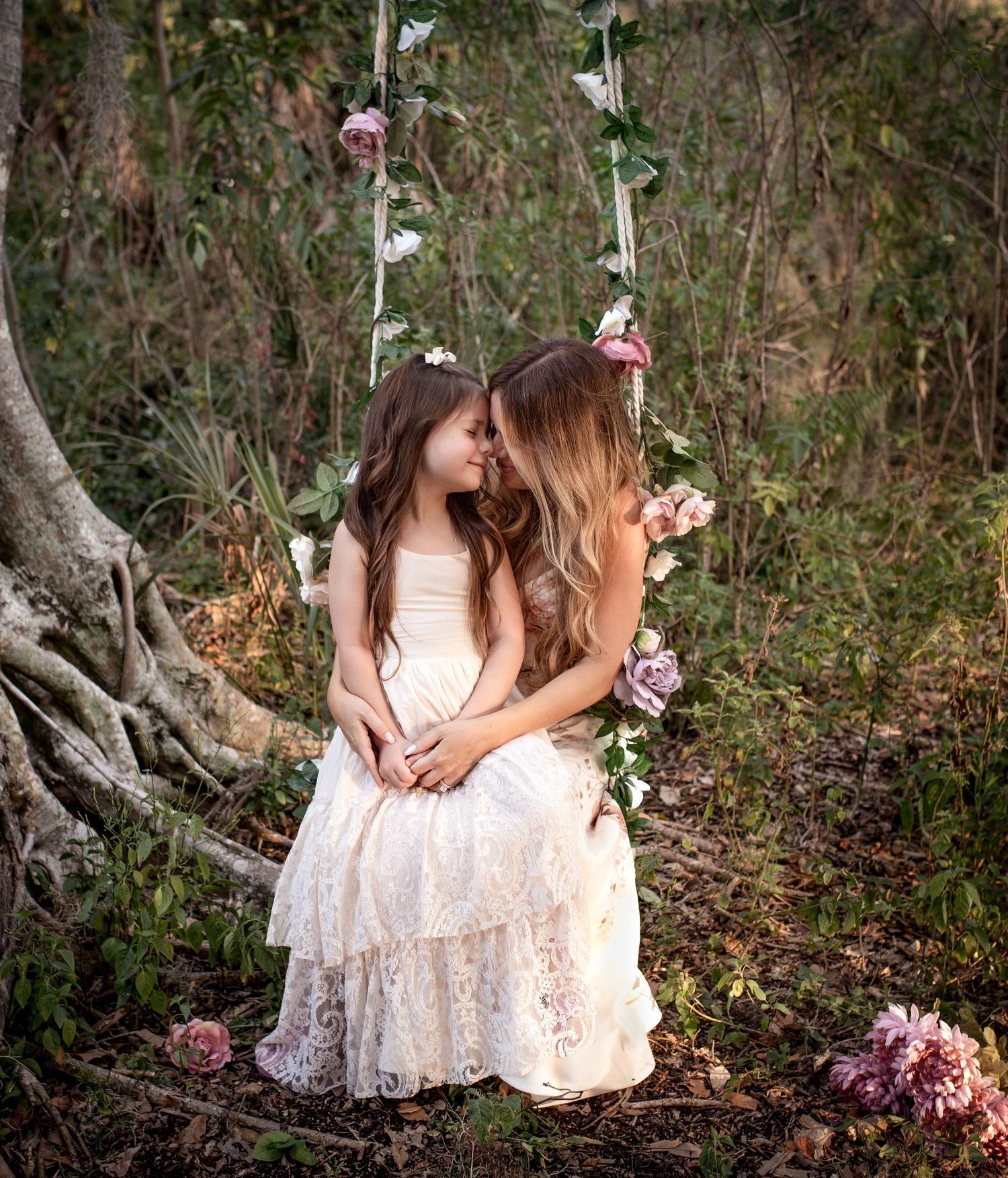 A girl in a white lace dress sits on her mom's lap on a flowery swing in outdoor setting with pink flowers nearby.