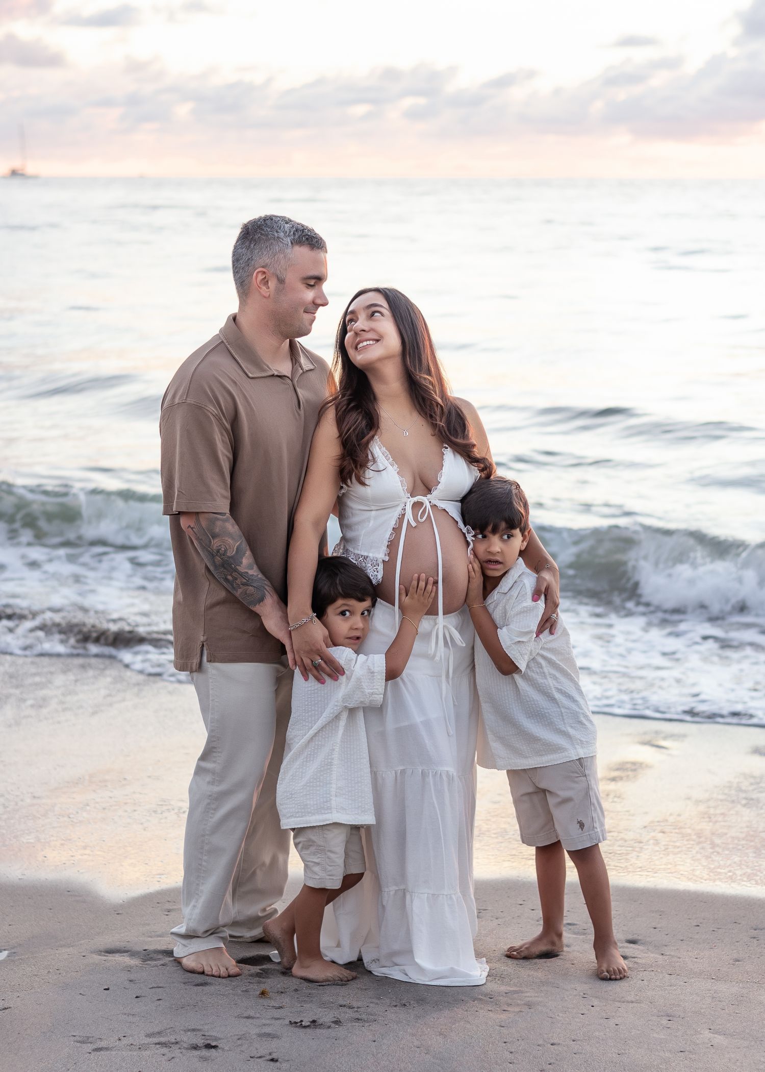 Family enjoying a tender moment together while walking along a sandy beach at sunset with ocean waves in background.