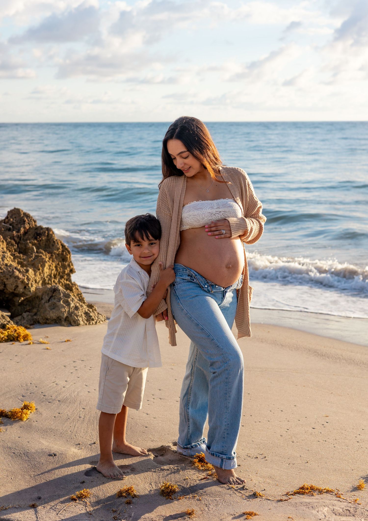 A tender moment captured at the beach as a child in white dress touches a pregnant woman's belly in the golden sunlight.