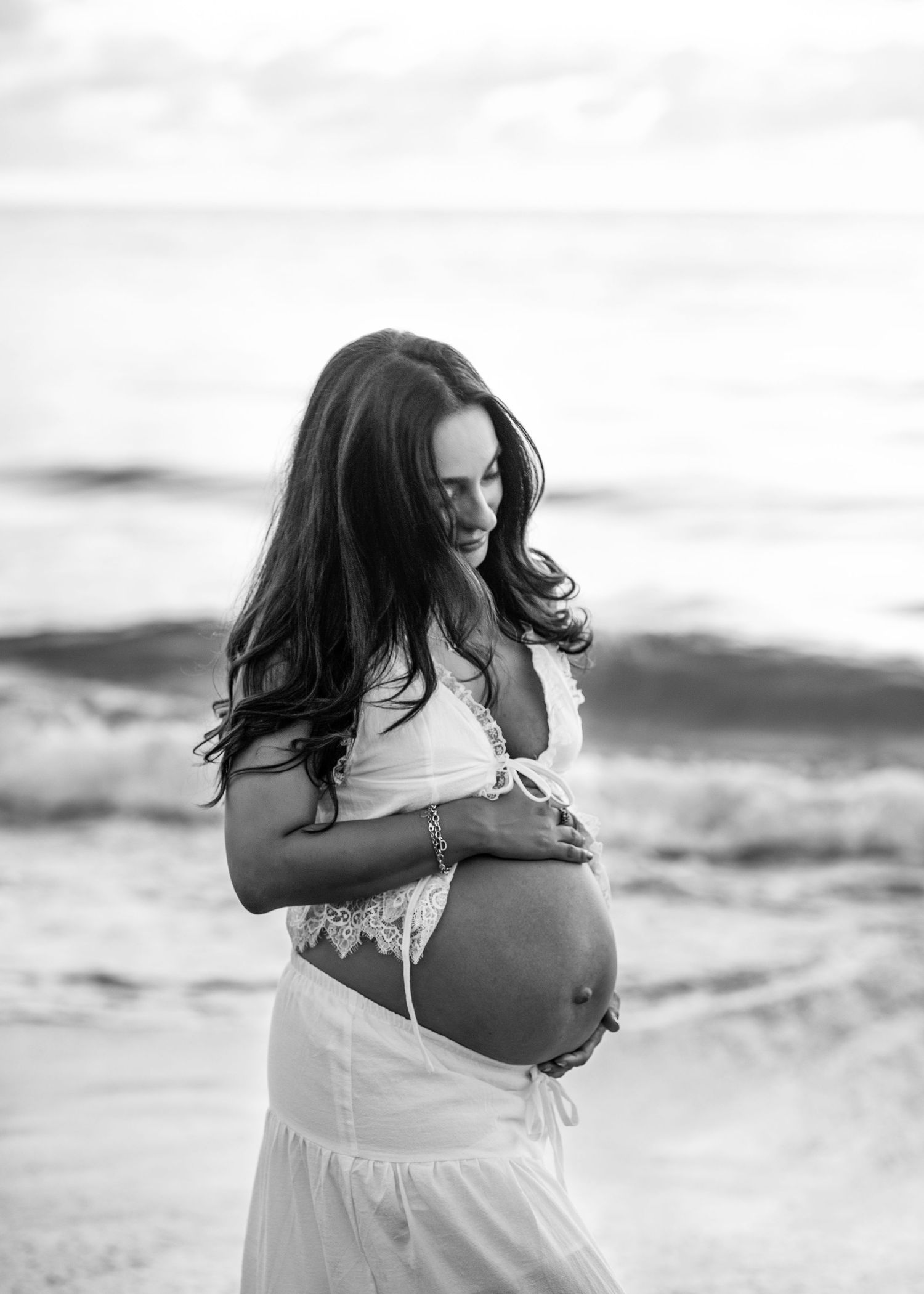 Black and white maternity photo series showing woman in white dress cradling baby bump on beach at sunset.