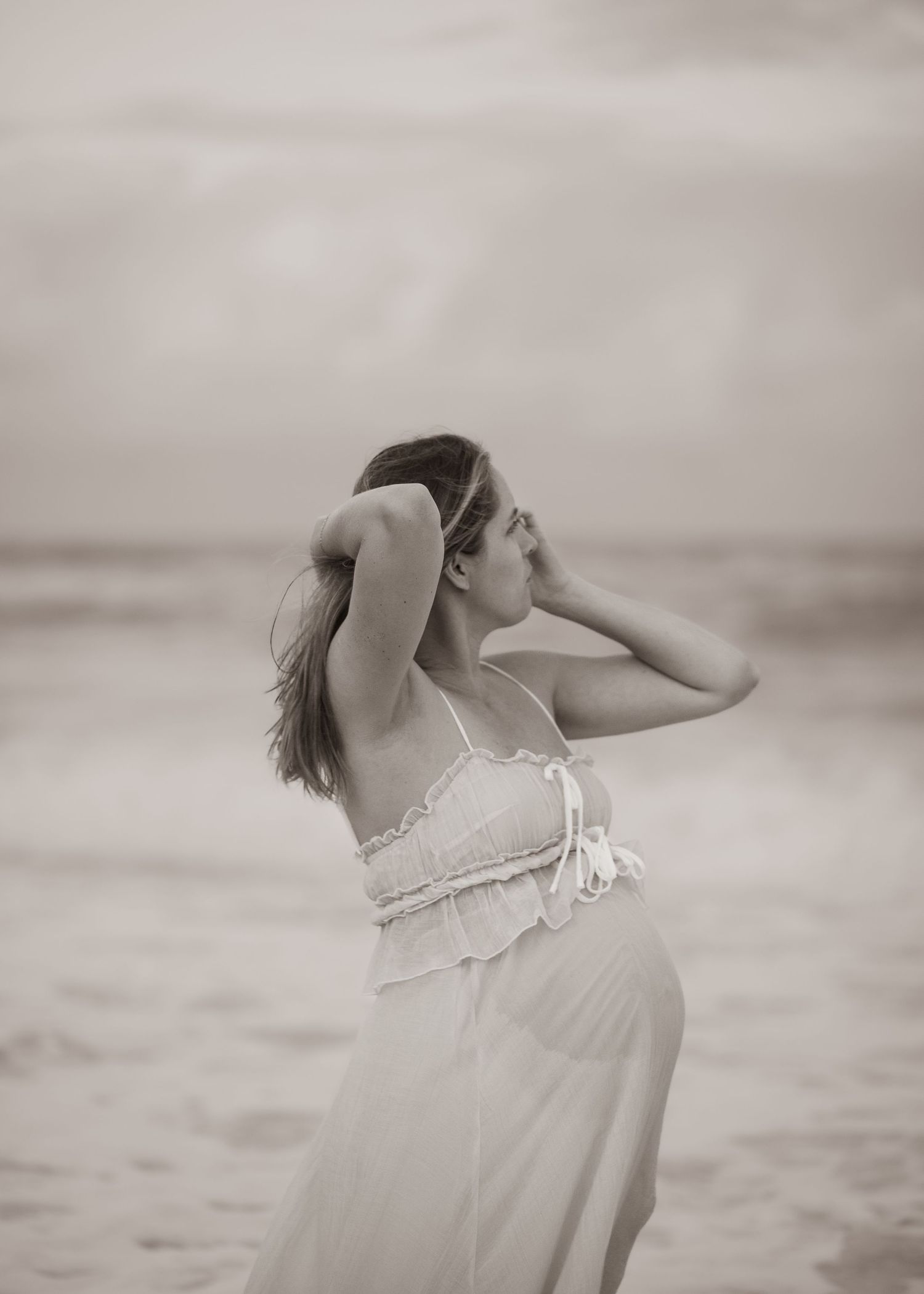 A black and white artistic maternity photo of an expectant mother in a flowing white dress posing on the beach.