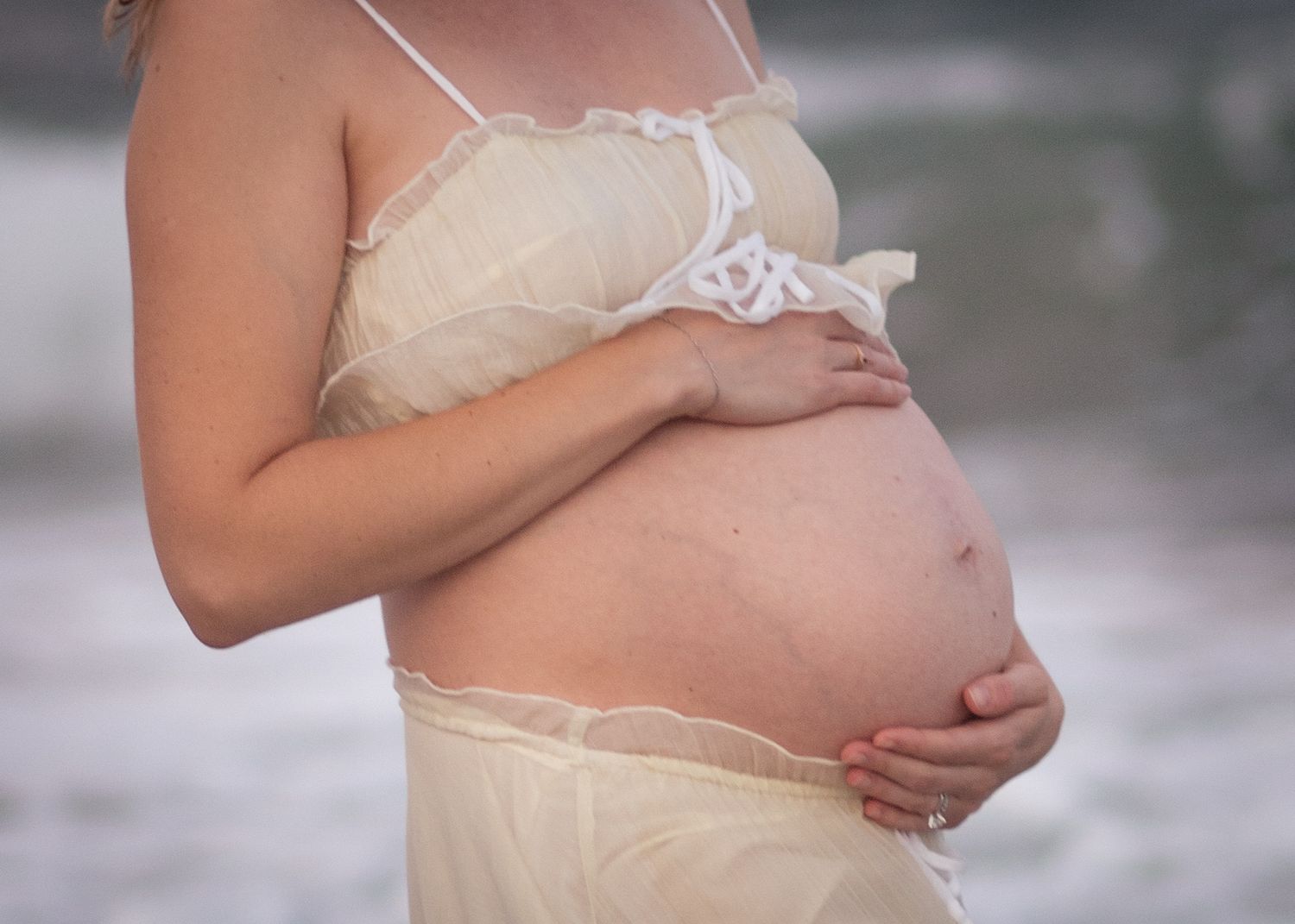 A pregnant woman in white lace lingerie gently cradles her baby bump in a soft-focus portrait.