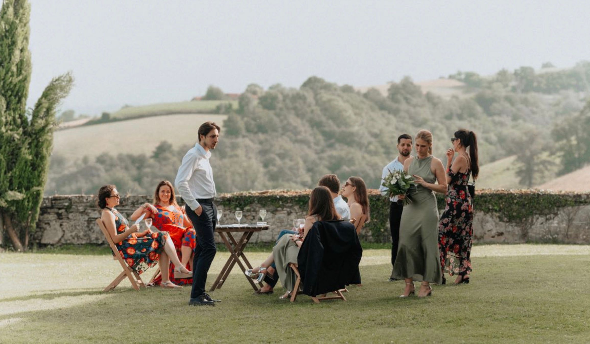 A group of people gathered on a grassy lawn with rolling hills in the background during an outdoor event.
