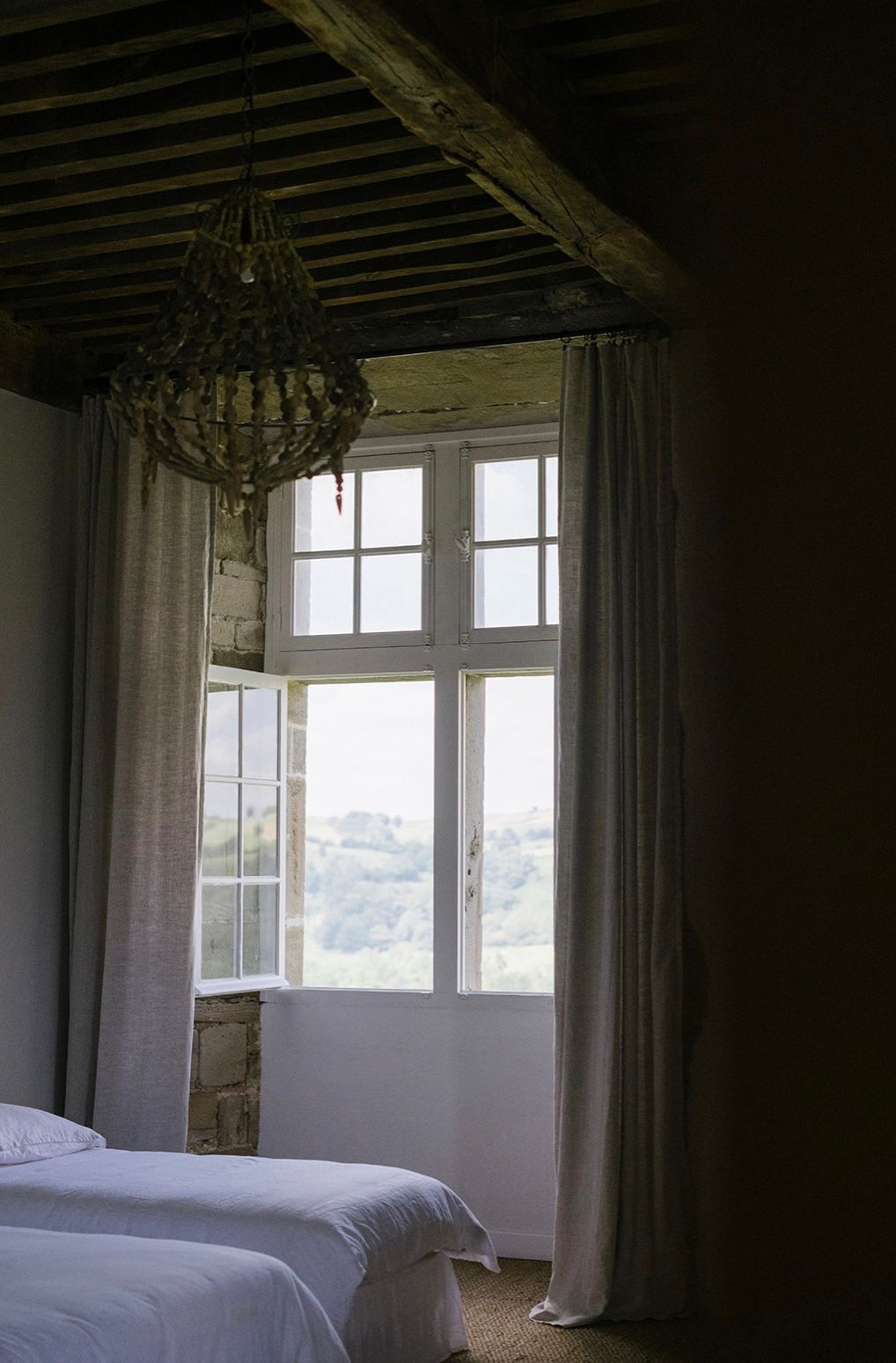 A rustic bedroom with a large window, white curtains and an ornate chandelier hanging from exposed wooden beams.