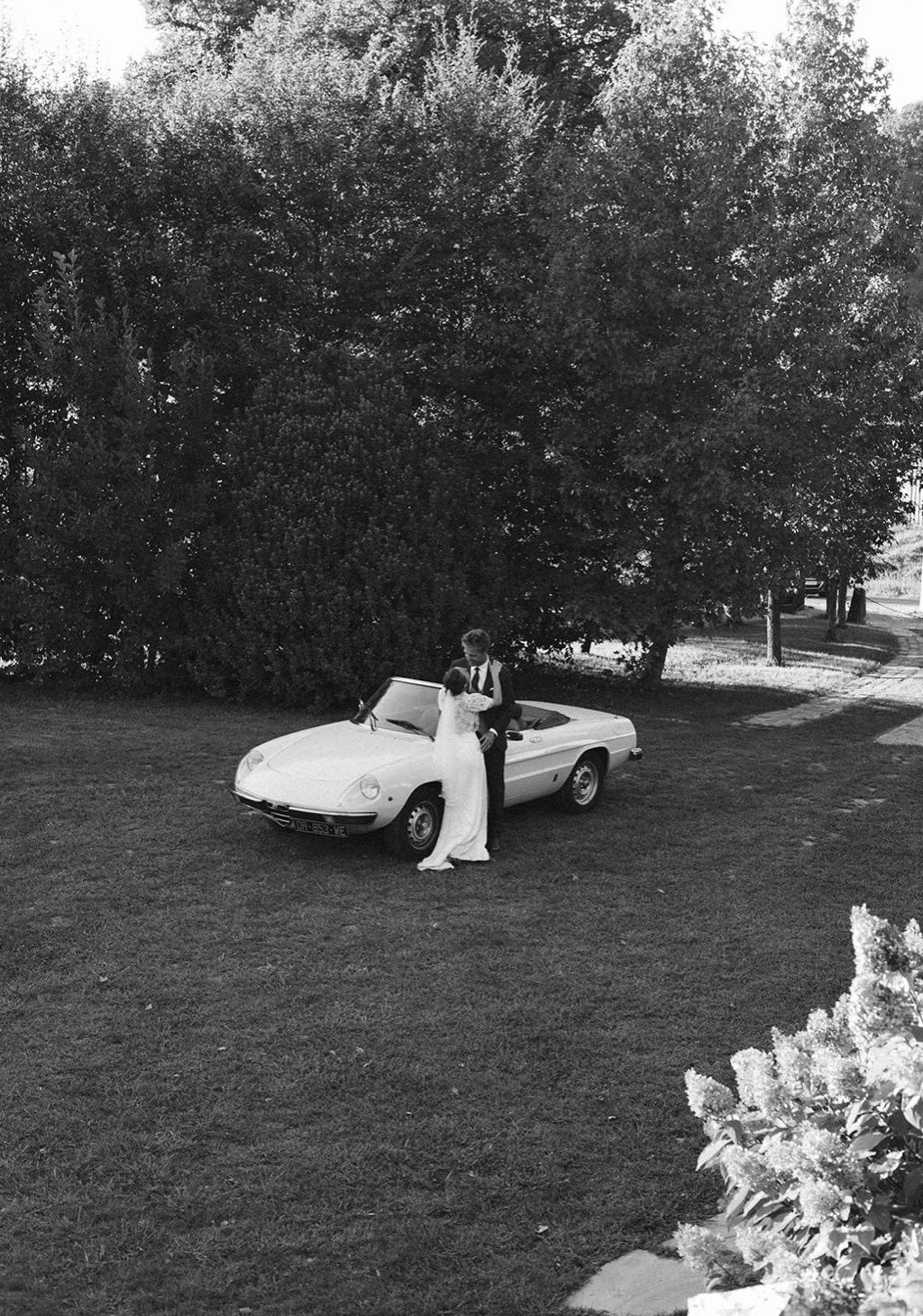 A series of black and white photographs showing a classic convertible car parked on a grassy lawn near bushes and trees.