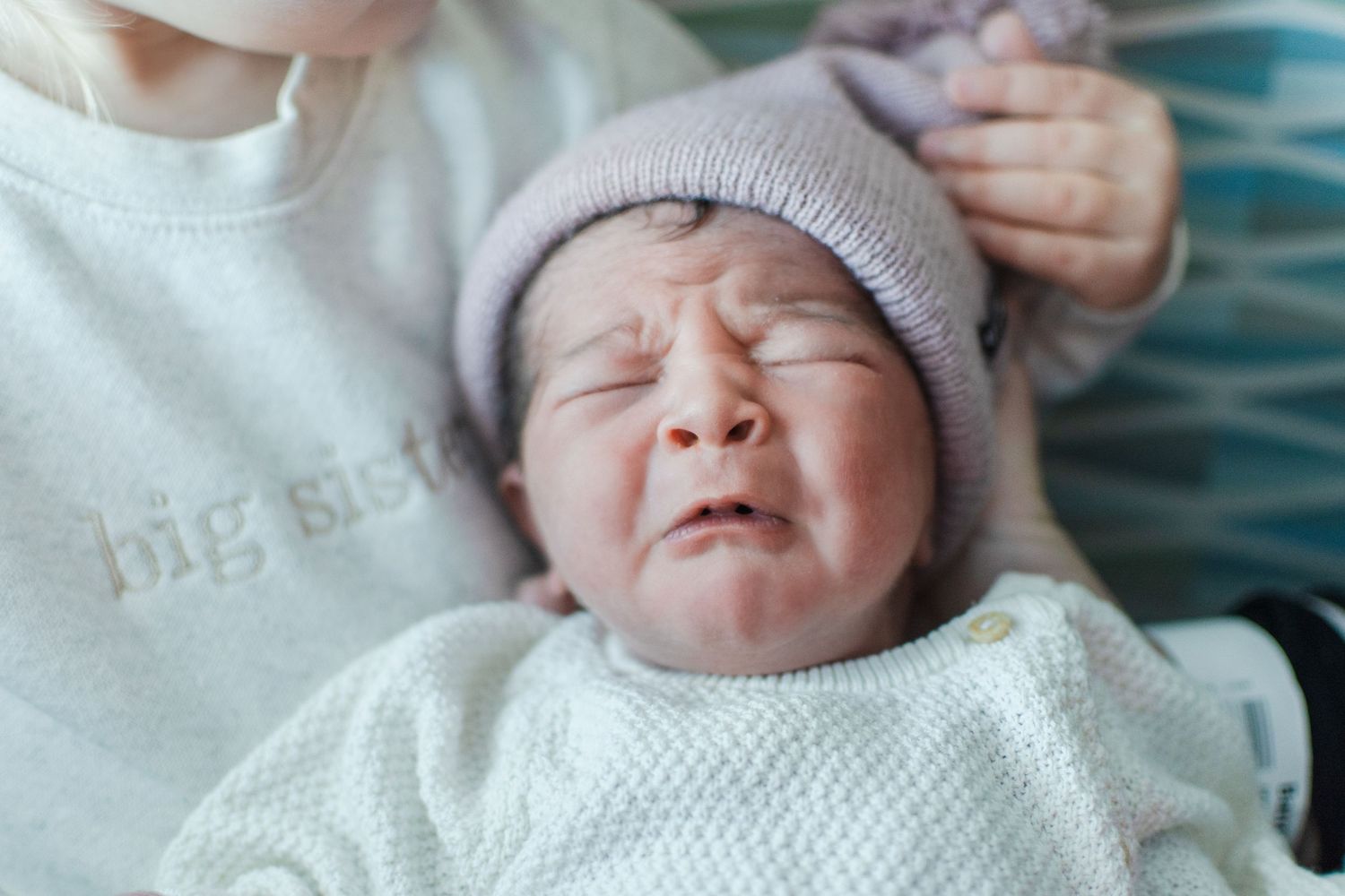 Crying newborn wearing pink knit hat and white sweater.