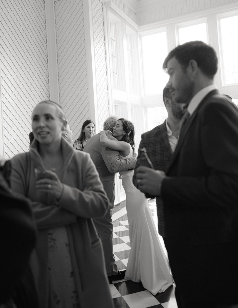 A candid black and white photo of wedding guests mingling in a bright indoor space.