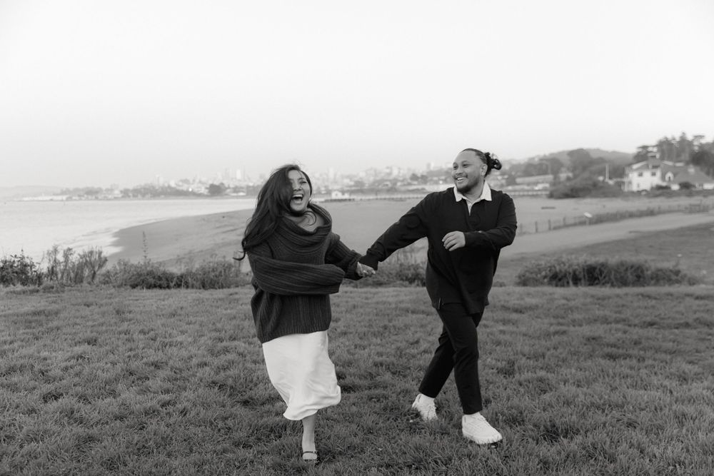 Black and white photo of two people running playfully through a grassy hillside.