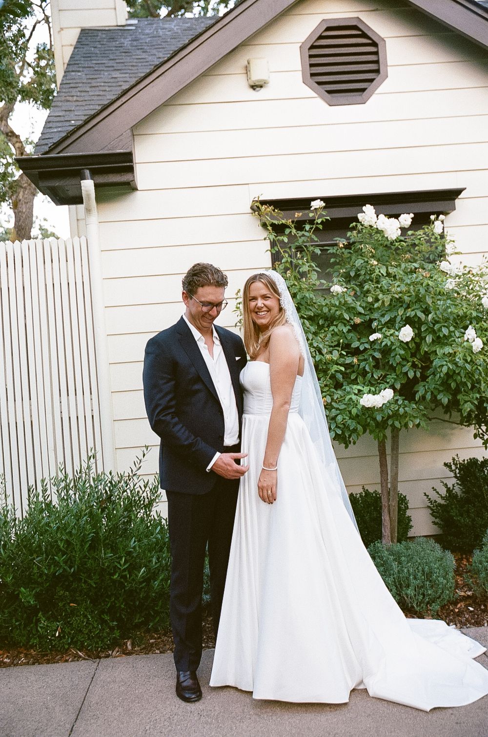 A couple poses in front of a white house with a picket fence and garden on their wedding day.