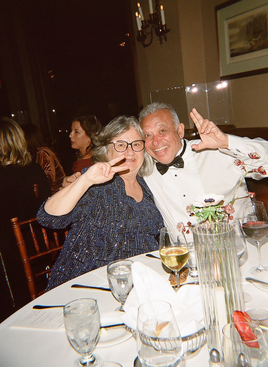 Two people making silly faces at a formal dinner party with wine glasses and table settings visible.