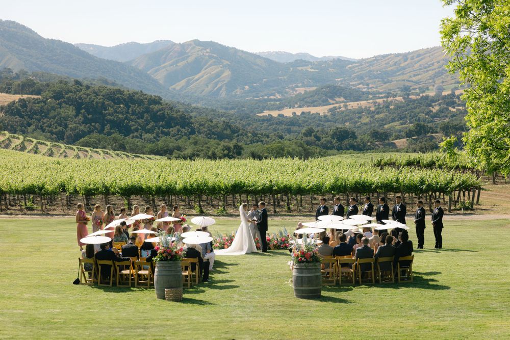 An outdoor wedding ceremony setup with rows of chairs on a grassy lawn overlooking vineyard hills and mountains.