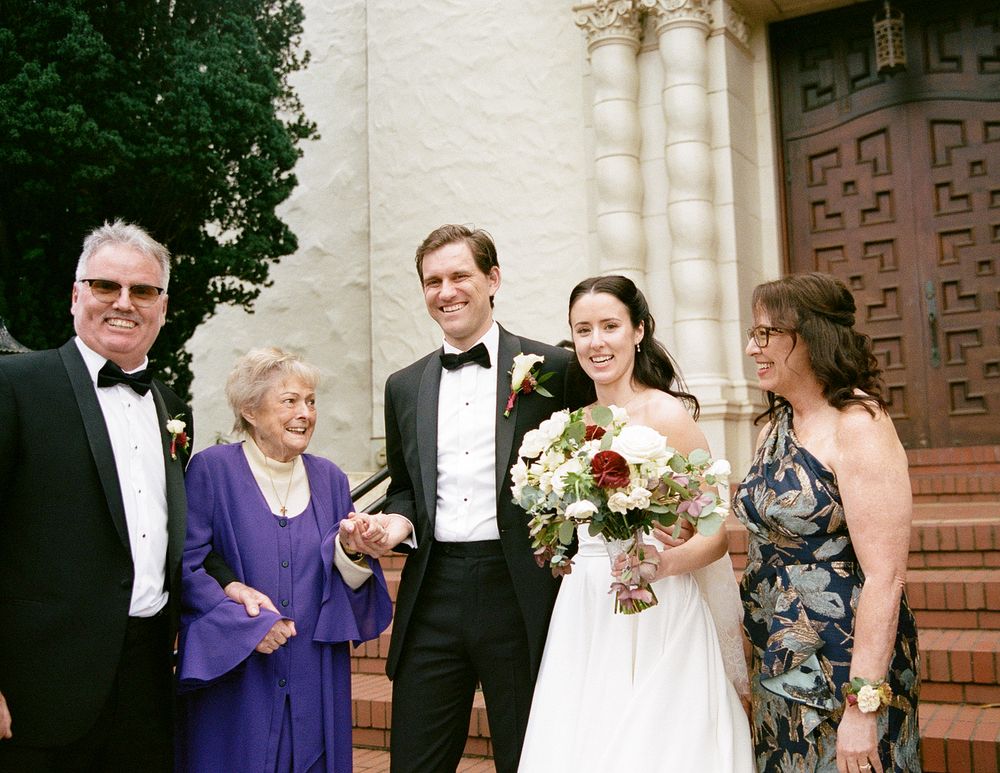Wedding party members stand together outside a church entrance with ornate wooden doors and stone columns.