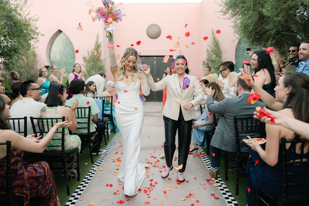 Newlyweds walking down aisle while guests throw rose petals during outdoor ceremony exit.