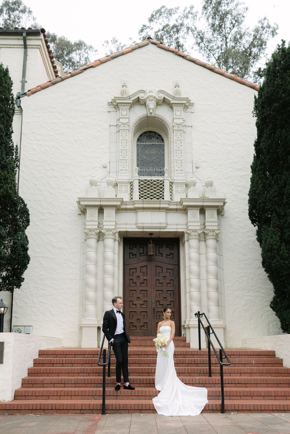 Couple posing on steps of a Spanish colonial style church with white stucco walls and ornate doorway.