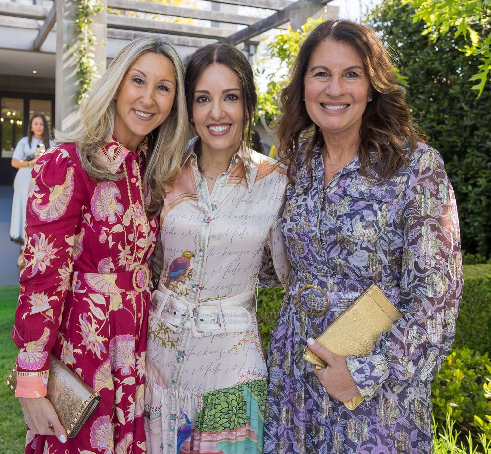 Three women pose together outdoors wearing floral print dresses in burgundy, white and purple tones.