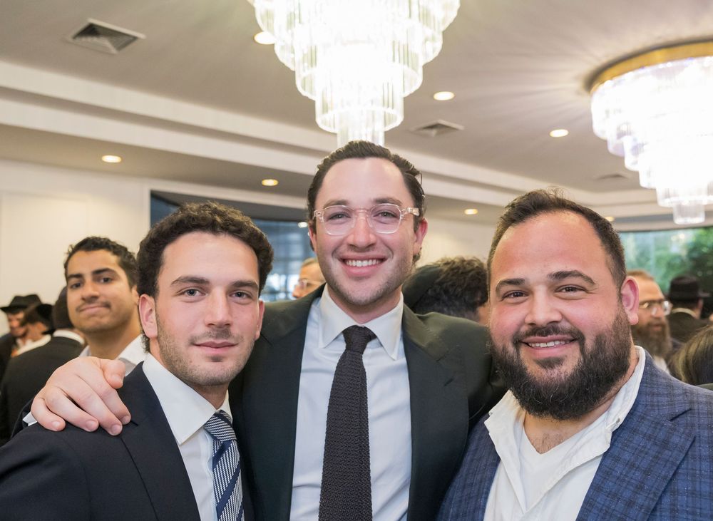 A group of men in formal attire and suits smile together at an indoor event with crystal chandeliers overhead.