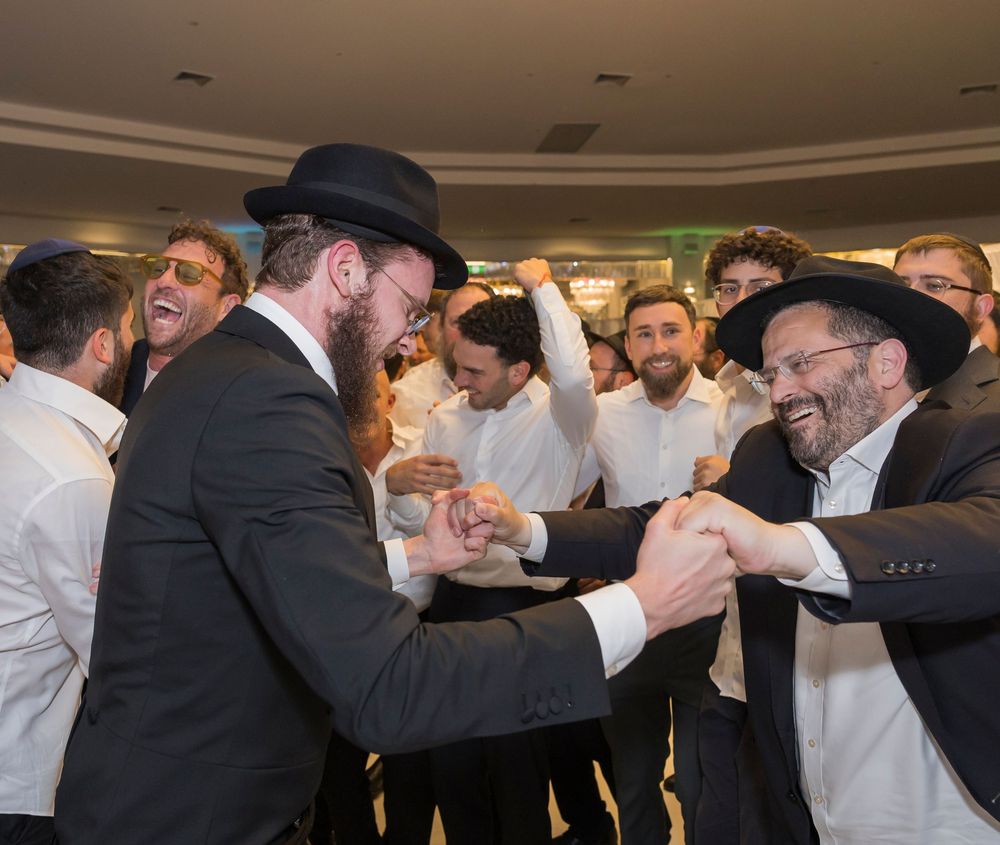 A group of Orthodox Jewish men dance and celebrate together in a joyous circle during a traditional gathering.