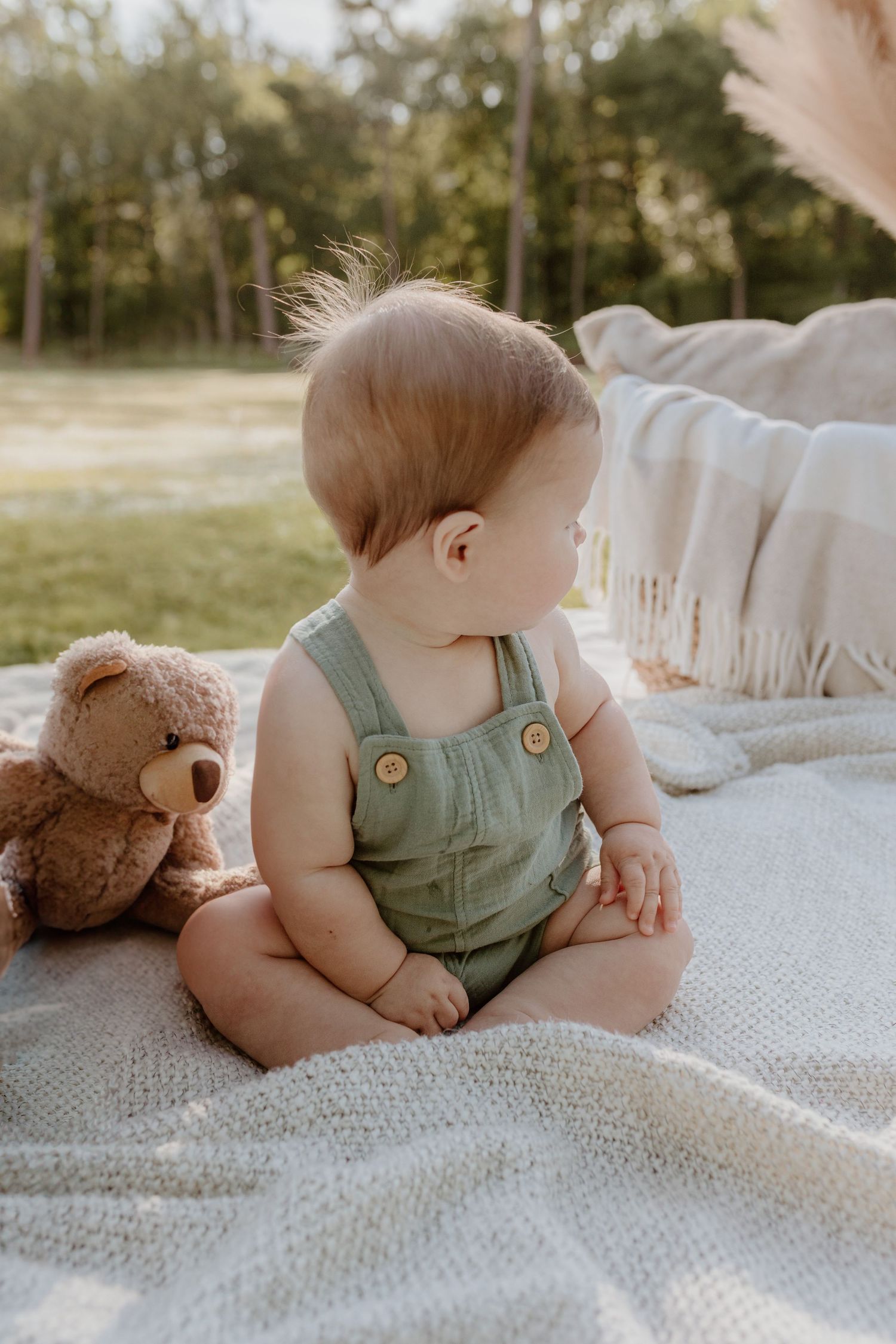 Baby in green overalls sits on white blanket with teddy bear during outdoor photoshoot.