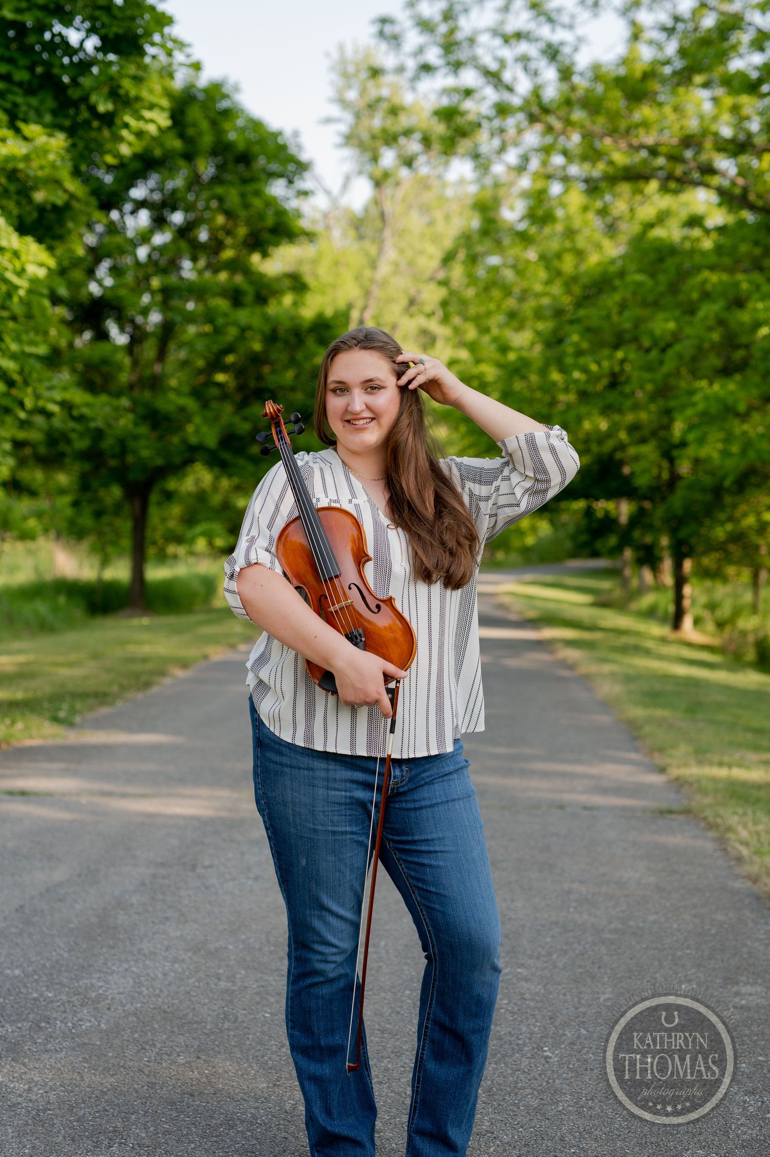 Musician in striped shirt holds violin while standing on tree-lined path.
