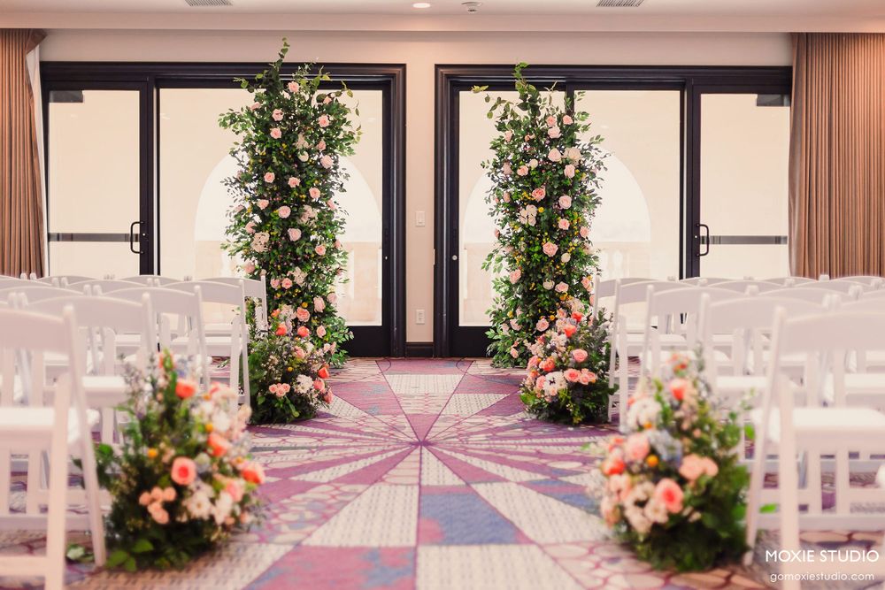 Indoor wedding ceremony setup with floral arches and white chairs on a geometric patterned carpet.