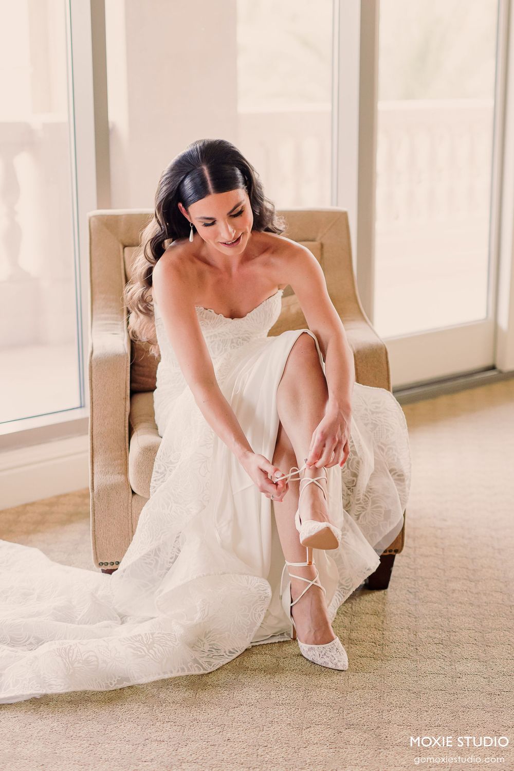 A bride in a white strapless wedding gown sits in a beige chair putting on elegant white strappy heels.