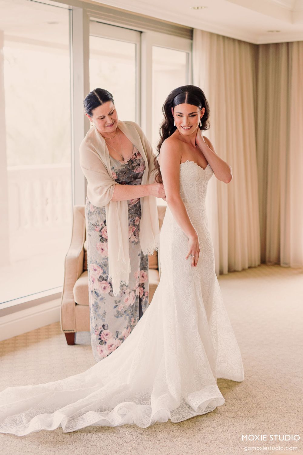 A bride in a fitted white wedding dress gets help with final adjustments in a sunlit room.