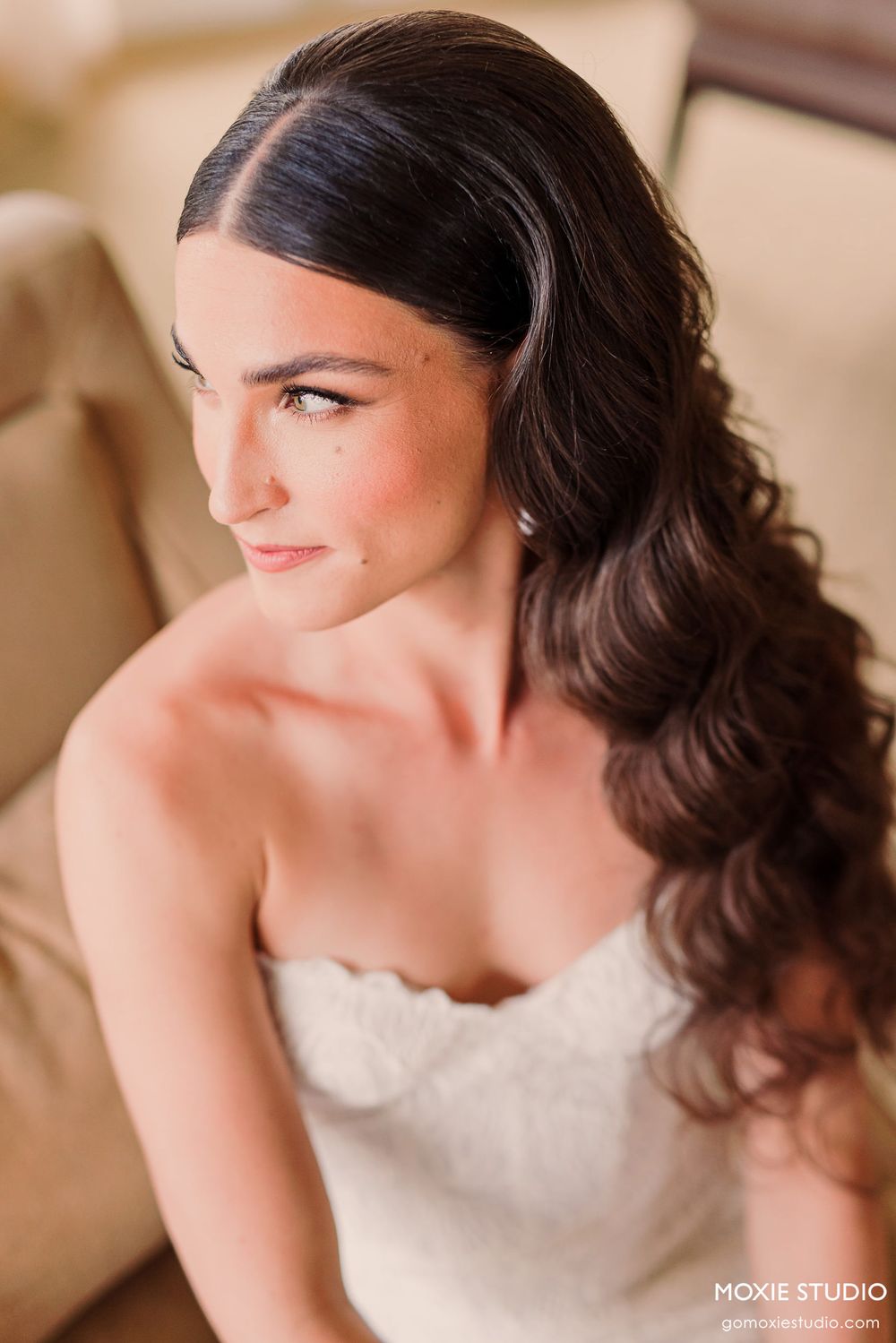 A bride in a white strapless gown with dark wavy hair styled elegantly looks to the side in a romantic portrait.