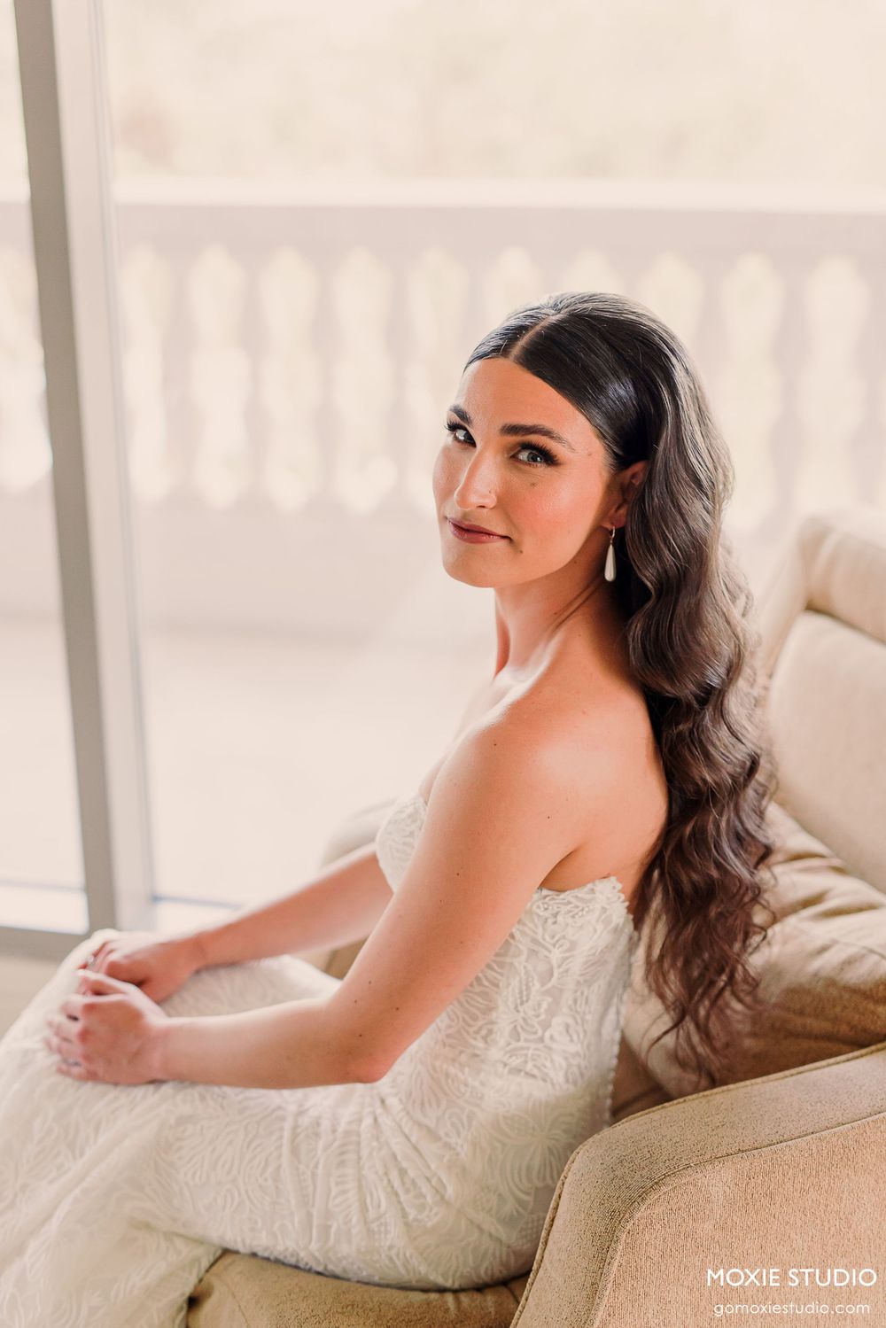 Bride in white lace gown lounges elegantly on beige sofa in romantic boudoir photoshoot.