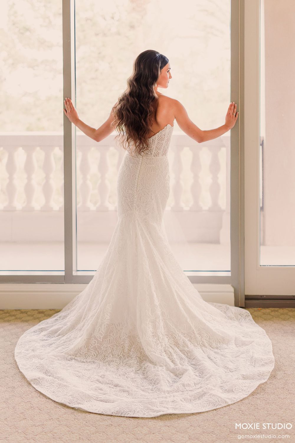 Elegant white wedding dress with long lace train displayed in a bright sunlit room with ornate balustrade.