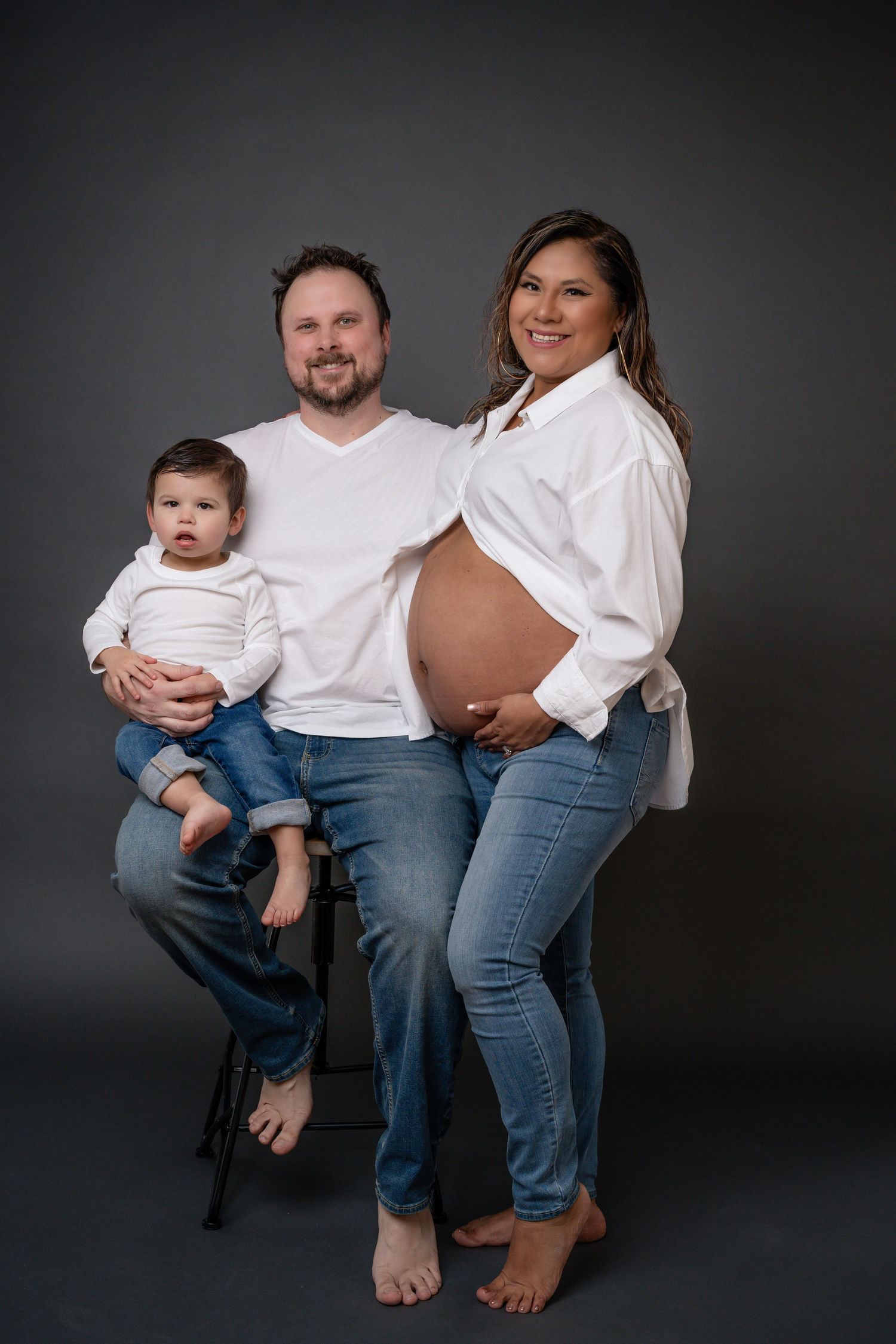 Family in matching white shirts and jeans posing casually against dark studio backdrop.
