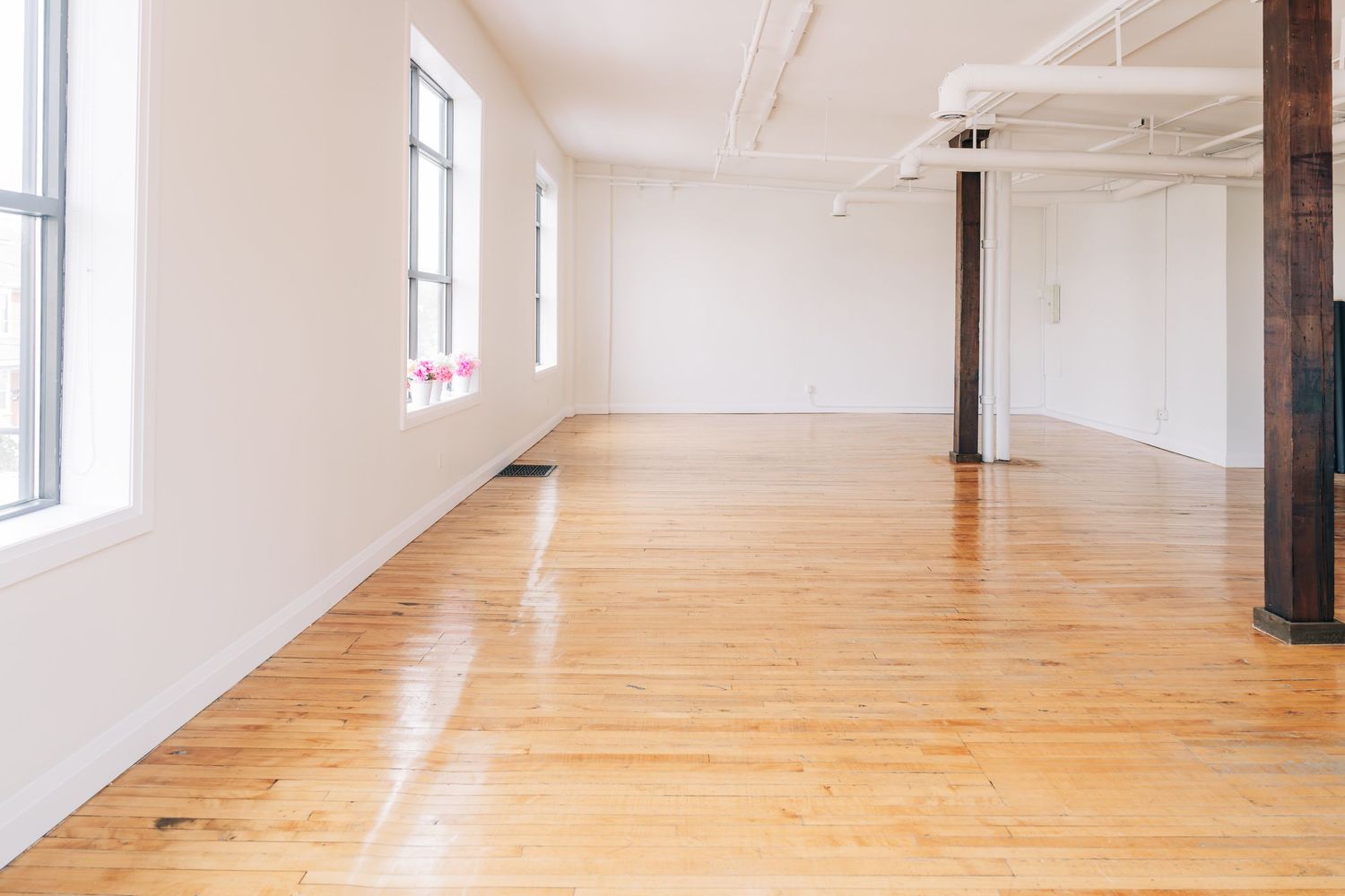 An empty bright studio space with polished hardwood floors, white walls, tall windows and exposed support columns.
